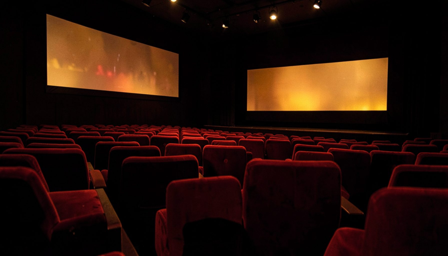 Dark atmospheric cinema interior with rows of red velvet seats and a glowing large screen, warm amber and deep crimson tones
