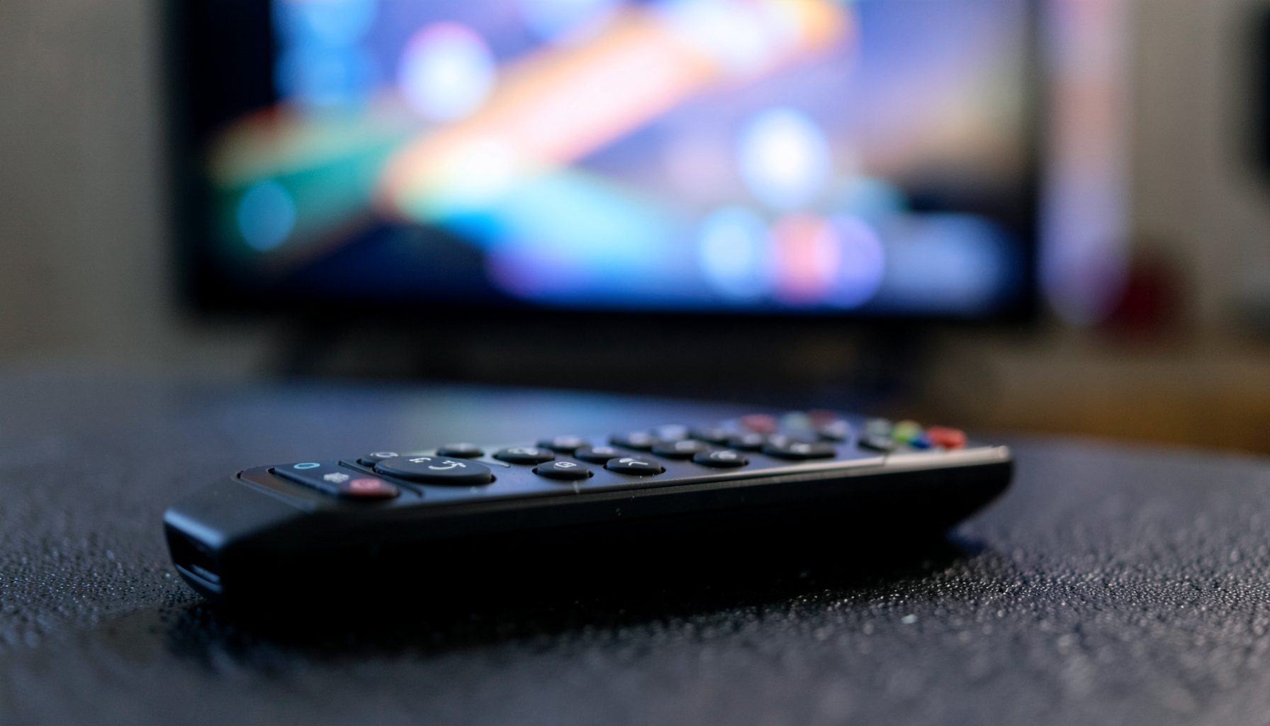 Close-up of a sleek remote control resting on a dark textured surface, with a softly blurred television screen glowing in the background