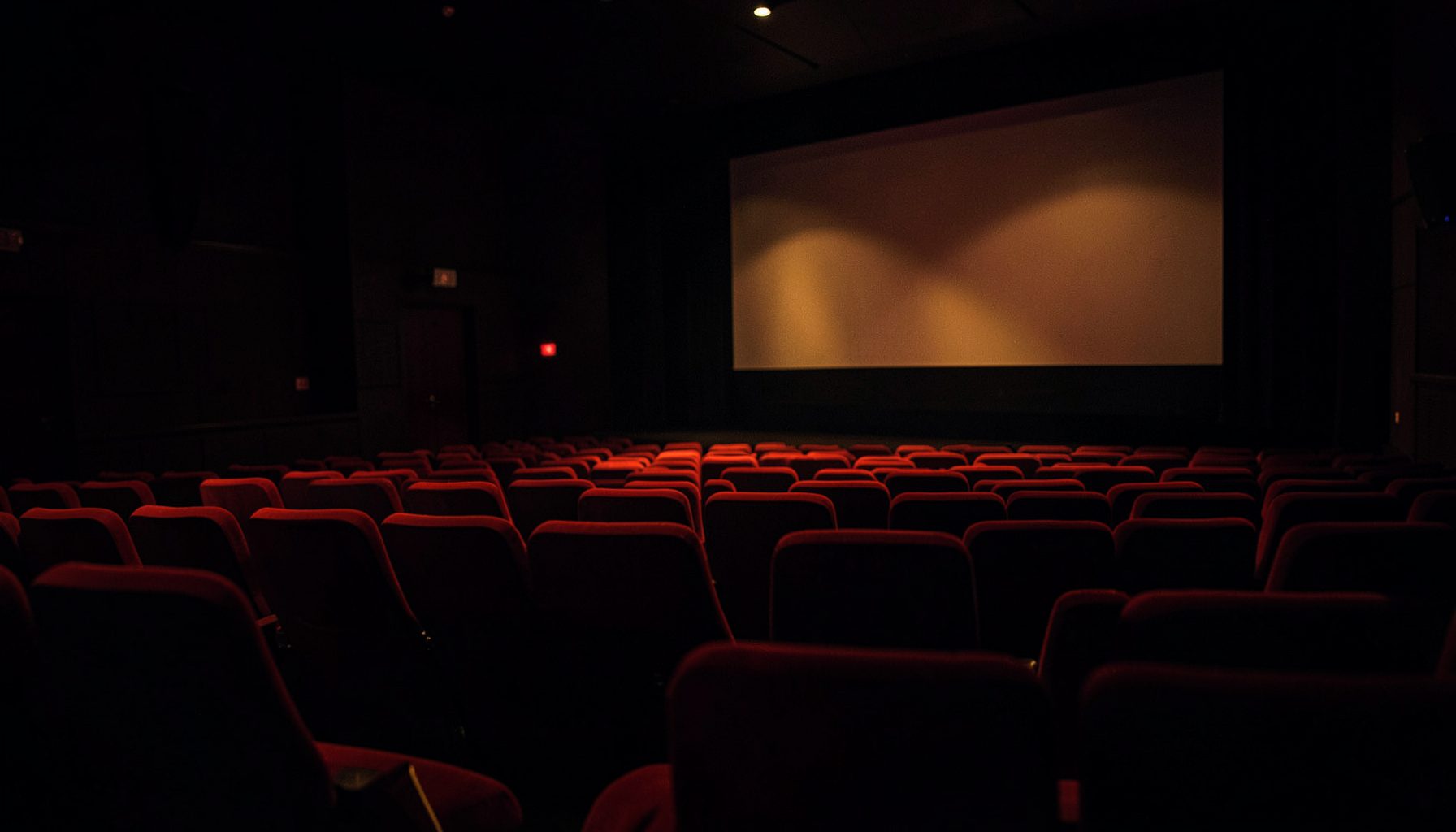 Dark cinema interior with rows of empty red velvet seats, soft ambient glow from the screen casting warm light across the auditorium, moody and cinematic atmosphere