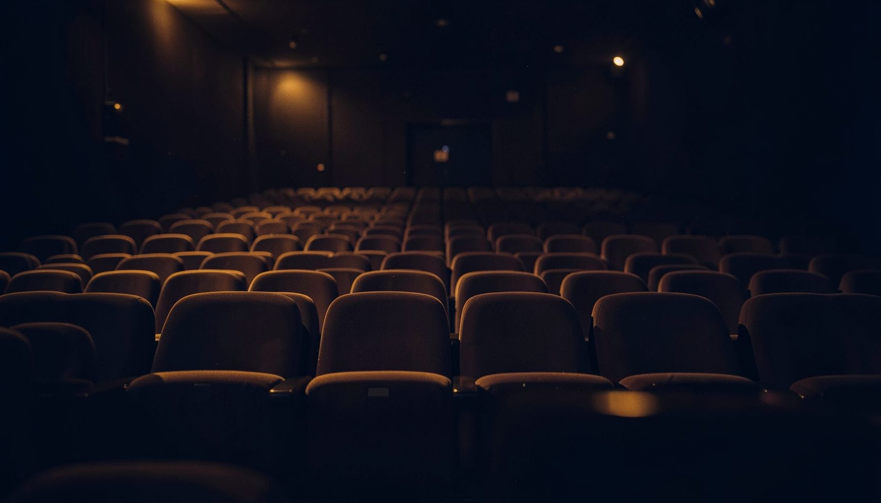 A wide cinematic collage of warm amber and deep navy tones, evoking the atmosphere of a darkened movie theater with soft light spilling across rows of seats